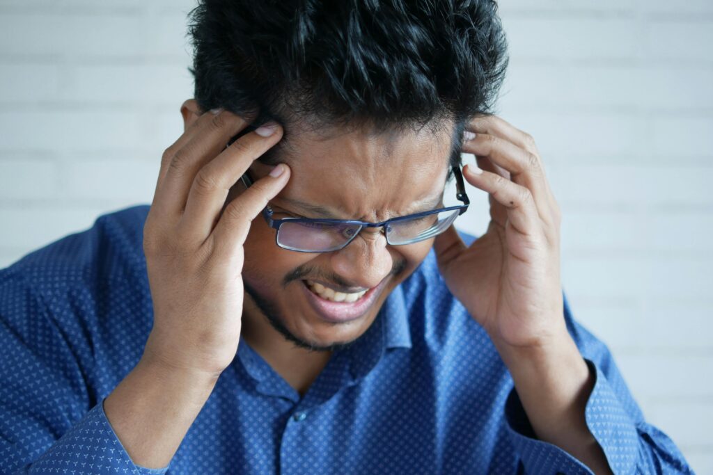 Close-up of man with eyeglasses and blue shirt experiencing a headache, showing frustration.
